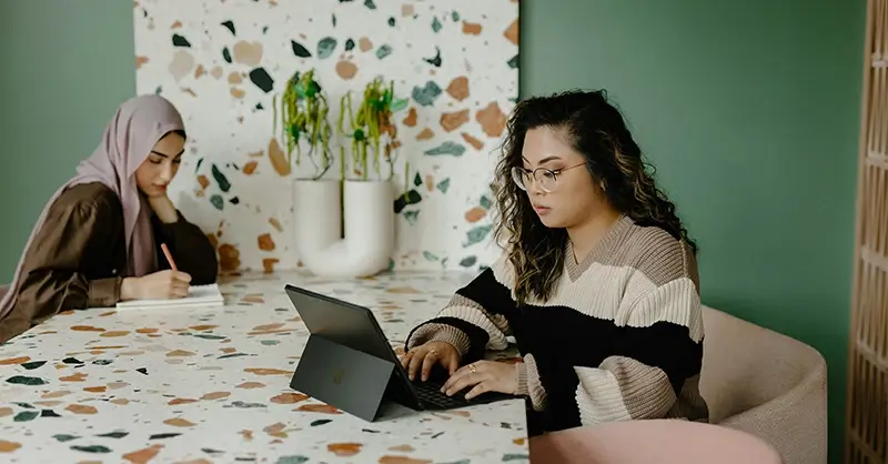 Women sitting together, one is on a computer while another writes notes