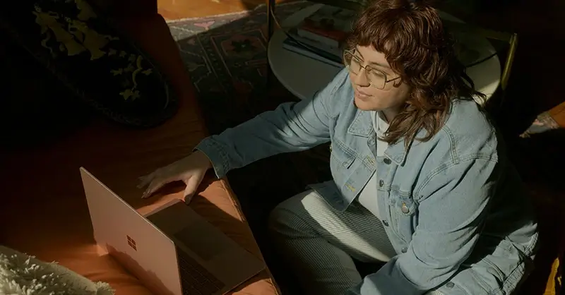A woman with glasses and a denim jacket on a video call on her laptop in her living room